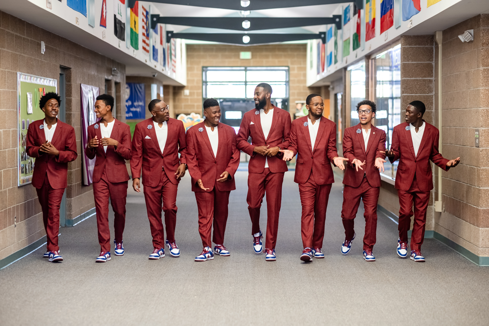 Photo of 7 MSU Denver MiSTERs and the Call Me MiSTER Director, Rashad Anderson, wearing matching suits, walking side by side down a hallway at a school.