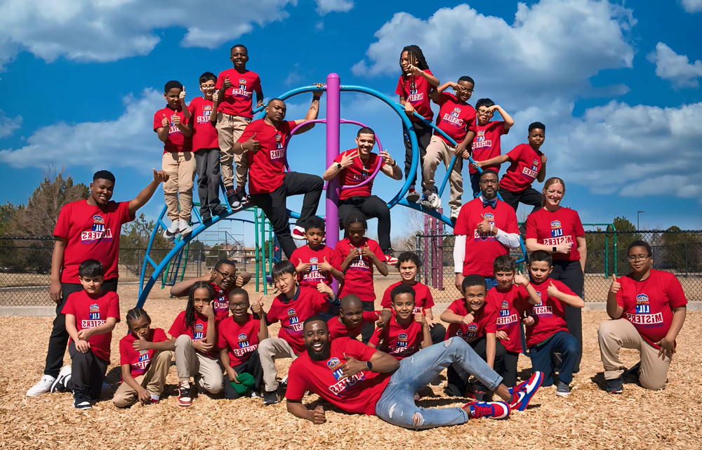 A photo of several MiSTERS posing on a piece of playground equipment with a group of young children. Everyone is wearing a red t-shirt with the text 