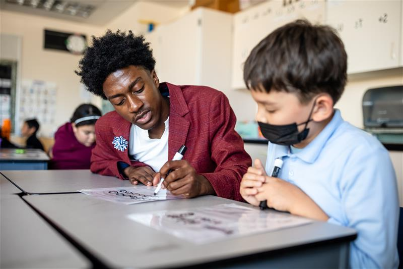 Photo of one of the MSU Denver MISTERs sitting with a young child at a desk in a classroom, writing on a piece of paper.