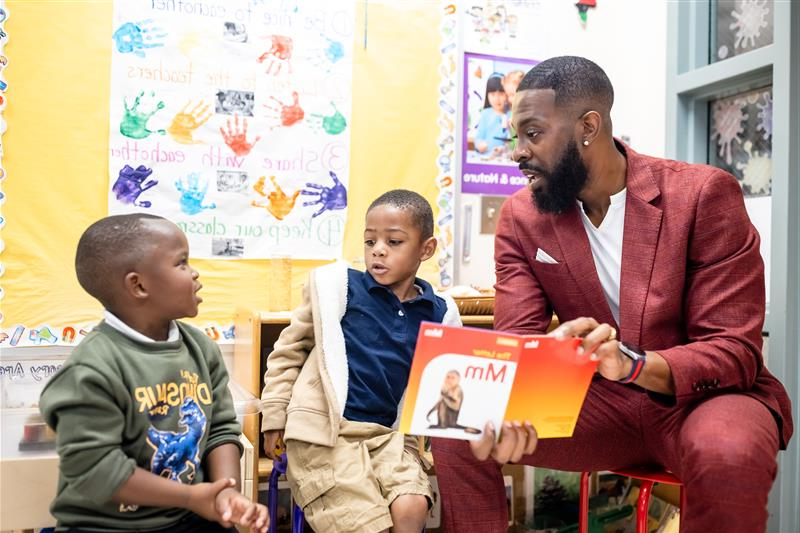 Dr. Rashad Anderson sitting with two young students in a classroom, reading them a book.